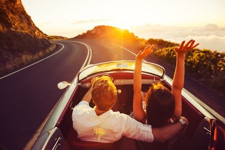 happy couple driving on country road into the sunset in classic vintage sports car